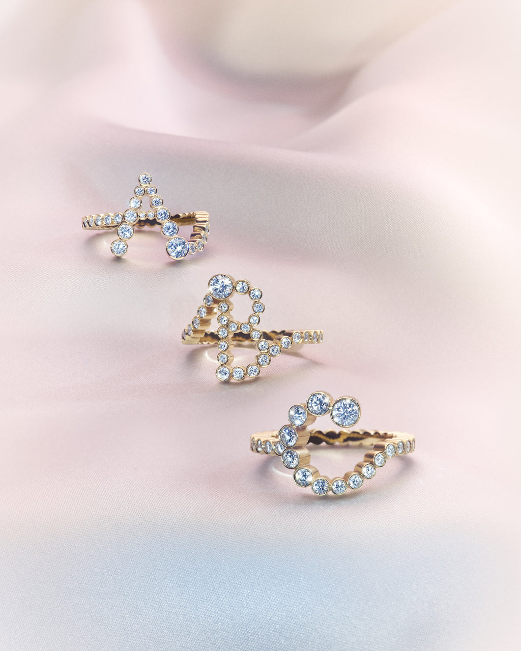 Three 18K yellow gold diamond rings shaped in the letters 'A', 'B' and 'C' on a soft pink background.